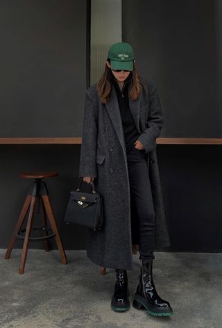 how to wear chelsea boots is shown in a photo of a woman standing in a cafe wearing a green baseball cap with a black coat over a black jumper styled with black skinny jeans and black chelsea boots