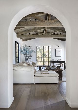 white living room through archway with wooden beamed ceiling and white sofas and fireplace with low round coffee table and crittal style windows
