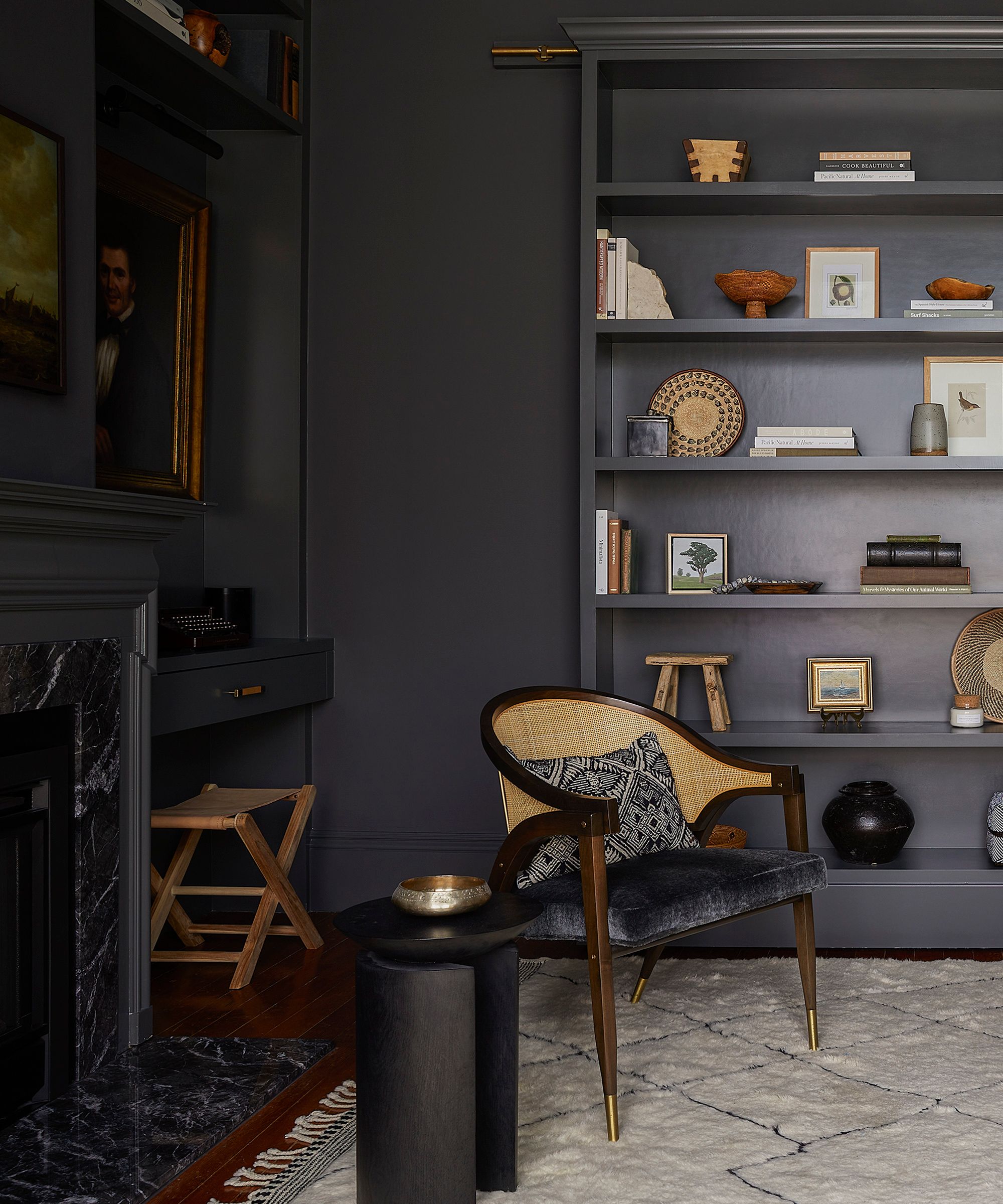 Tall, dark gray built-in bookshelf filled with books, baskets, and framed artwork, next to a cane-backed chair