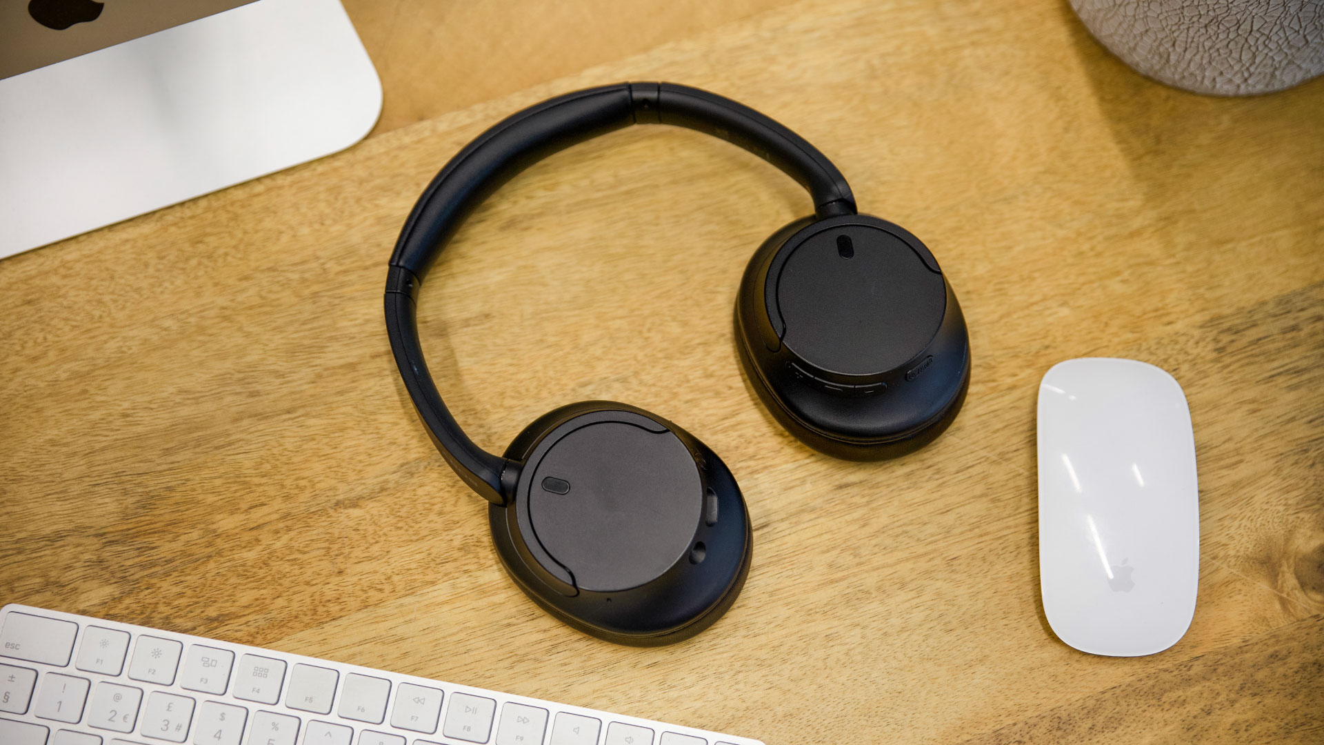 A pair of black over-ear headphones and a white computer mouse are centered on a wooden desk, with a white keyboard visible below them.
