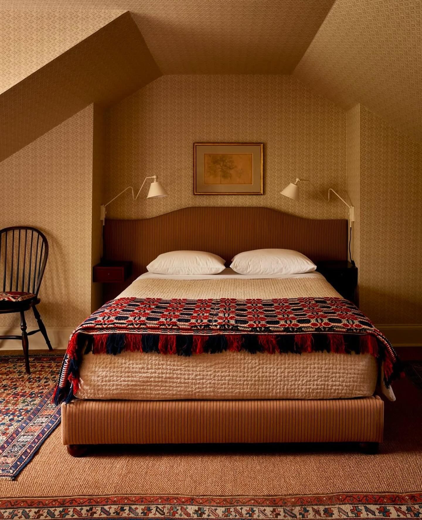 Image of a bedroom with caramel wallpaper and a bed that has a light brown fabric bedframe. There is cream bedding on the bed and two sconce lights on either bedside. 