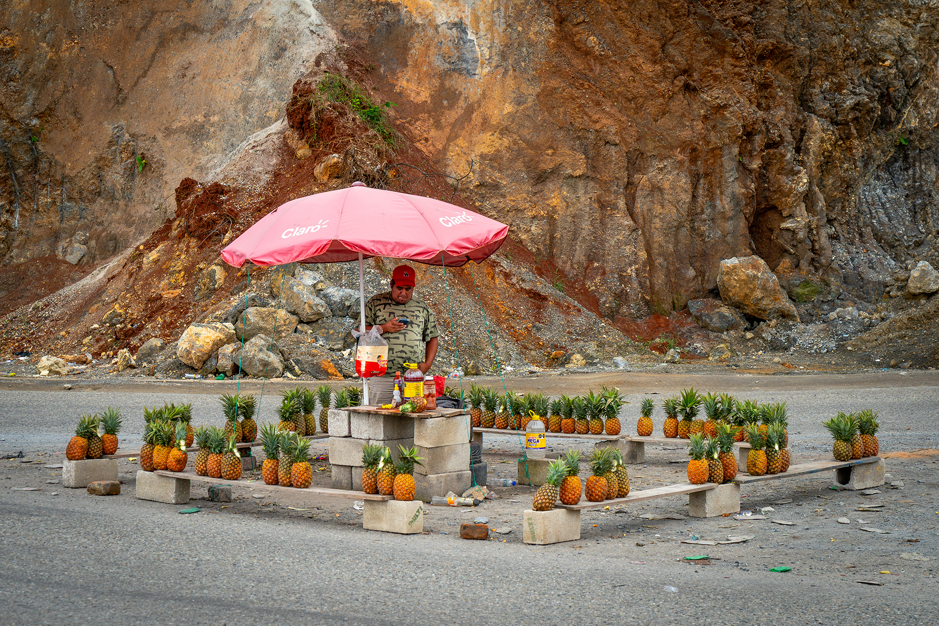A roadside pineapple stand with a man under a pink umbrella. The stand is made of cinder blocks, with pineapples neatly arranged in a rocky landscape