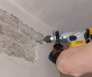 gloved hand holding drill drilling into brickwork where plaster has been removed