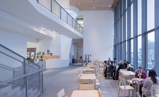 An open space in the building with white wall and floor to ceiling windows (divided with grey steel). by the windows are beech coloured tables and chairs which is partly occupied with people sitting. On the left of the seating area is a peek of the staircase and a lobby area