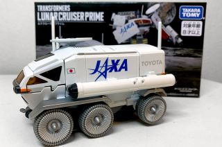closeup of a six-wheeled white moon rover transformer toy with the words "jaxa" and "toyota" written on its side, in front of the box it's sold in.
