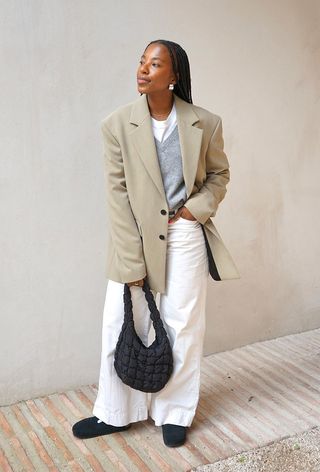 a photo showing what shoes to wear with wide-leg pants with a woman standing outside wearing a tan blazer over a gray sweater and white t-shirt styled with white wide leg jeans and black flat clog shoes