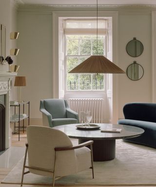 A white living room with a curved blue sofa, a circular wooden coffee table, a white fireplace and a low light fixture hanging from the ceiling