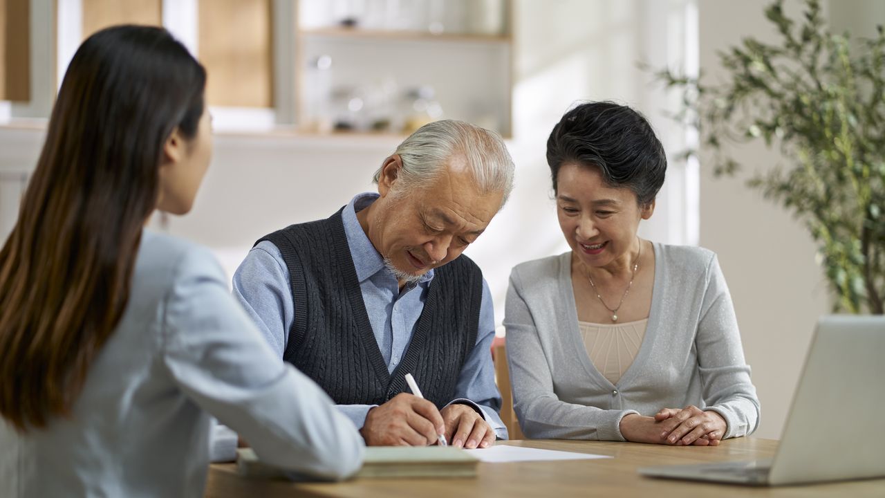 An older couple sign paperwork at a real estate agent&#039;s office.