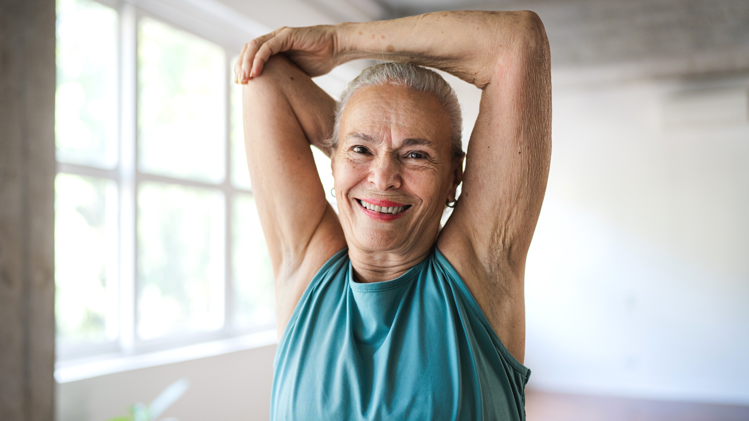 A senior woman stretches her arms above her head and smiles at the camera. 