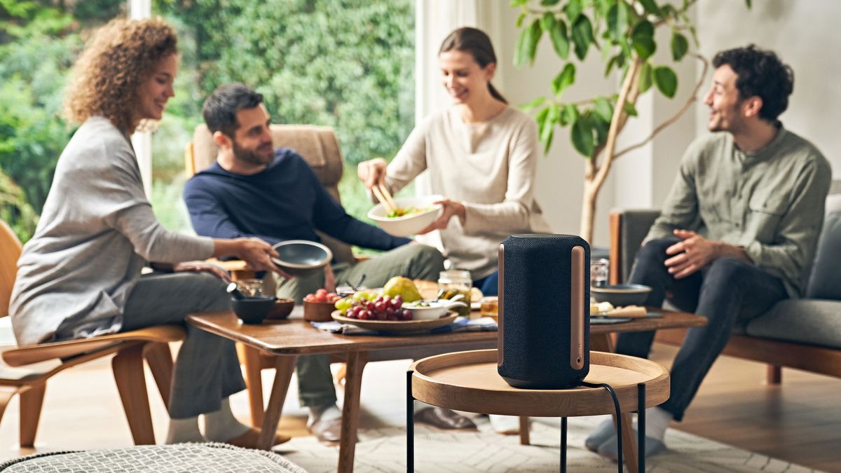 The Sony SRS-RA3000 wireless speaker sitting on a small round table, with a four people sitting in the background