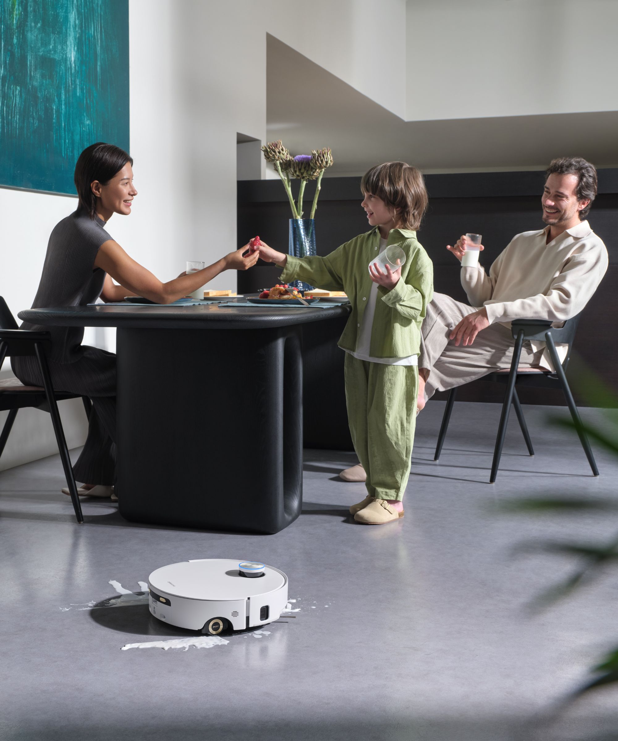 Dreame Aqua10 Ultra Roller on gray floor next to small milk puddle, pictured in front of happy family sharing food at a black table.