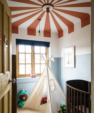 A pale blue, two-tone painted nursery with a red and white, big top-style, painted ceiling design and teepee.