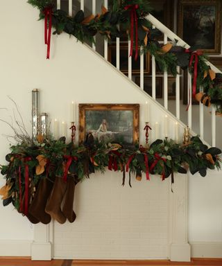 A white hallway decorated with a wreath draped over the stair banister decorated with red bows and layered with vintage gold candlesticks and vintage art