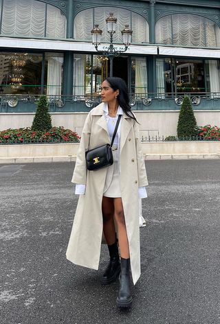 how to wear chelsea boots is shown in a photo of a woman walking down the street wearing a long tan trench coat over a white button-down shirt layered over a white tank top styled with a tan mini skirt, black crossbody bag, and black chelsea boots