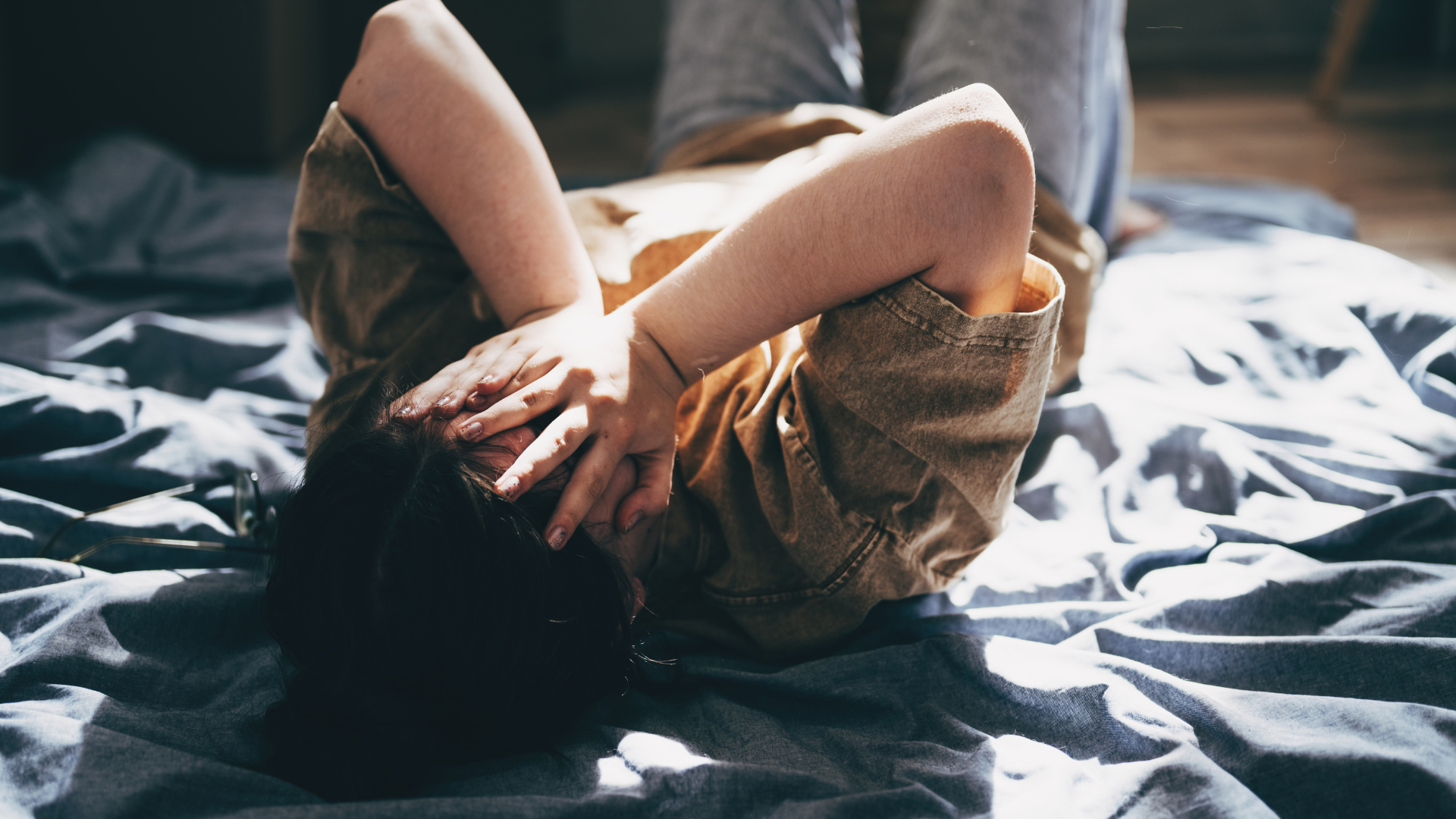 A teenager lies on their bed looking distressed with both hands covering their face.