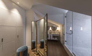 A hotel bathroom with a white sink counter, twin round mirrors, a large shower with a clear glass door, a large standing mirror, cupboards, a round seat and wooden floors.