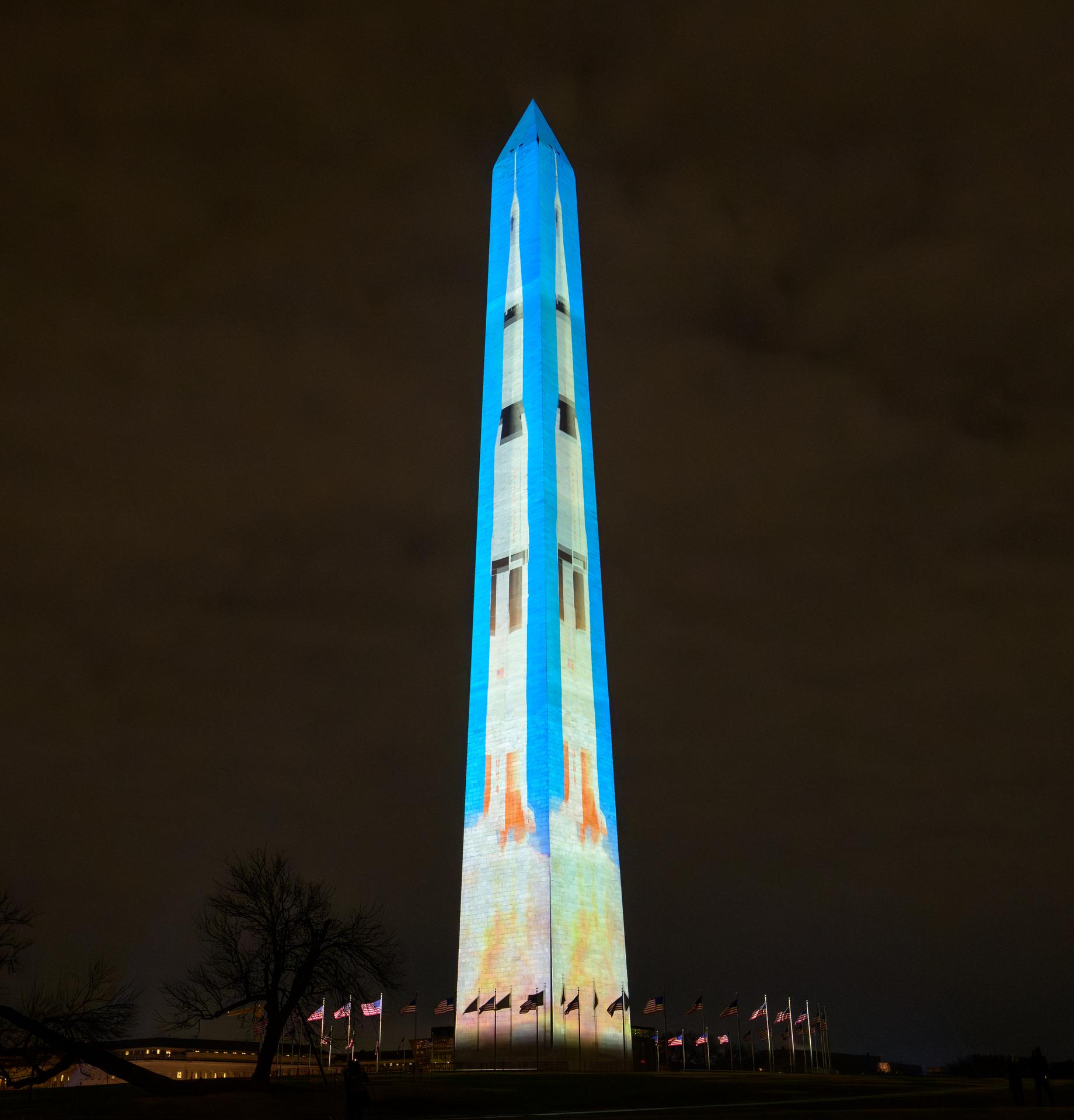 A tall obelisk-shaped monument is illuminated with projectors to produce scenes of rockets, astronauts on the moon, and the numbers 250 vertically down the building