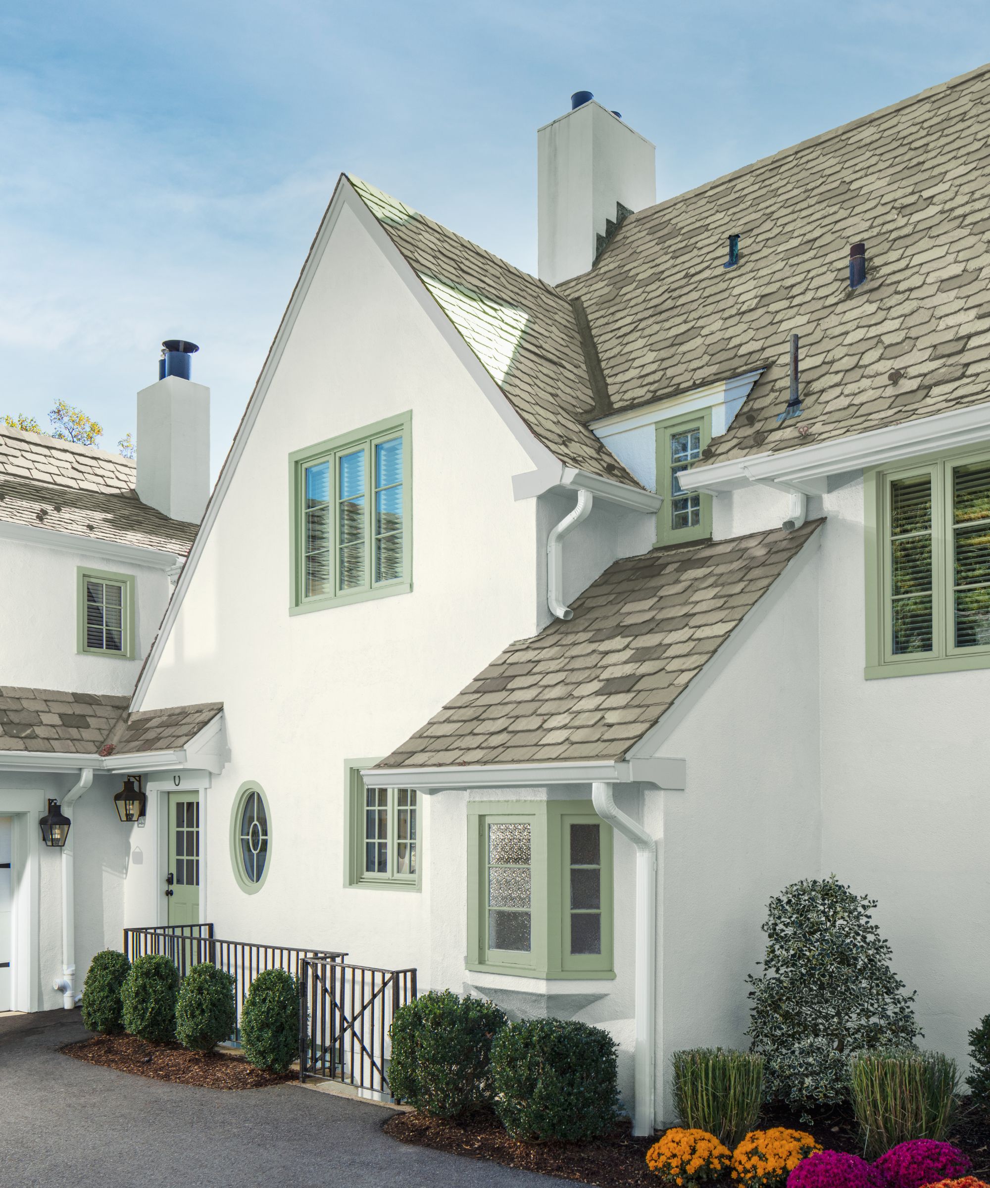 House with sage green painted window sills and Cloud White by Benjamin Moore painted exterior, grey slats on the roof, and hedges and colourful flowers surrounding it