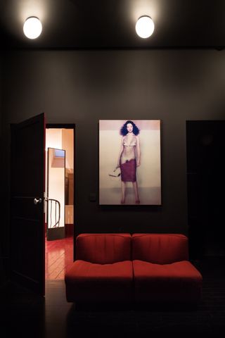 Dark painted room with dim lights and a red double couch and a painting of a woman on the wall