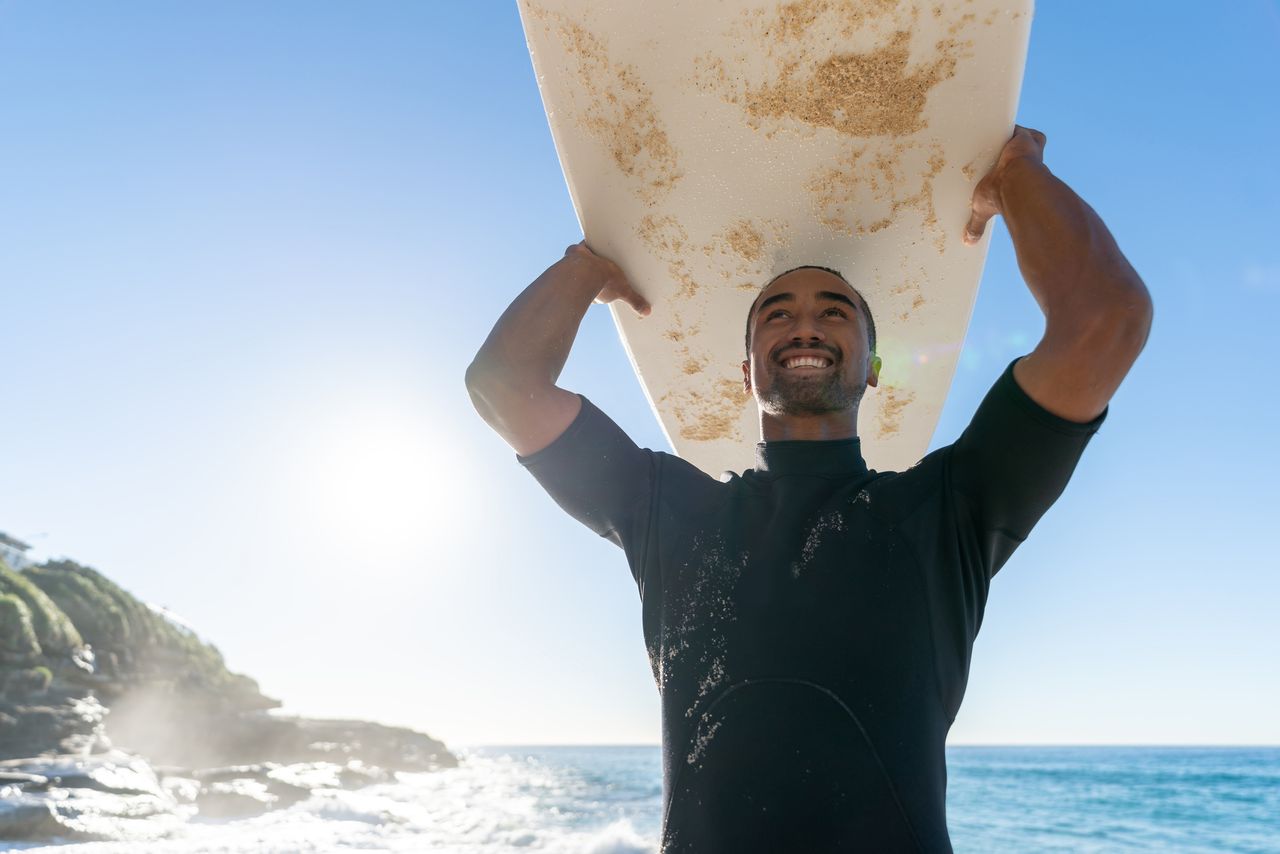 A man in his 40s holds a surfboard over his head and smiles.