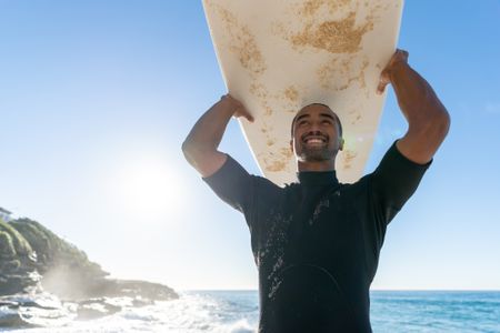 A man in his 40s holds a surfboard over his head and smiles.