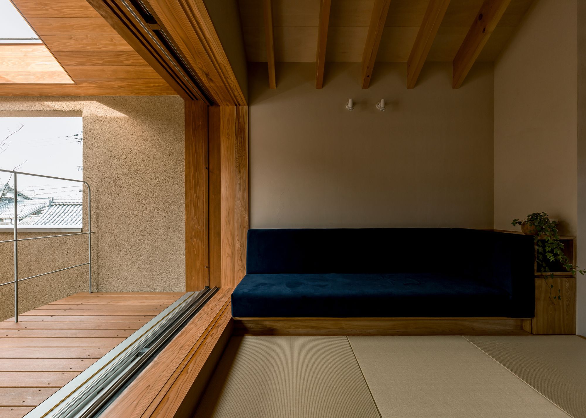 A minimalist indoor-outdoor Japanese living space with a built-in sofa on a wood frame with blue velvet material and one plant sits next to it