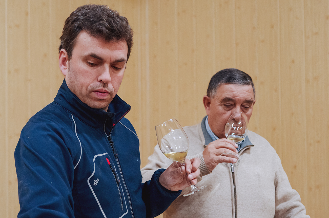 An&amp;iacute;bal Asensio Paunero and Jose Lorenzo Tejedor, both holding glasses of white wine to their noses, looking downwards, presumably towards a (off-screen) barrel