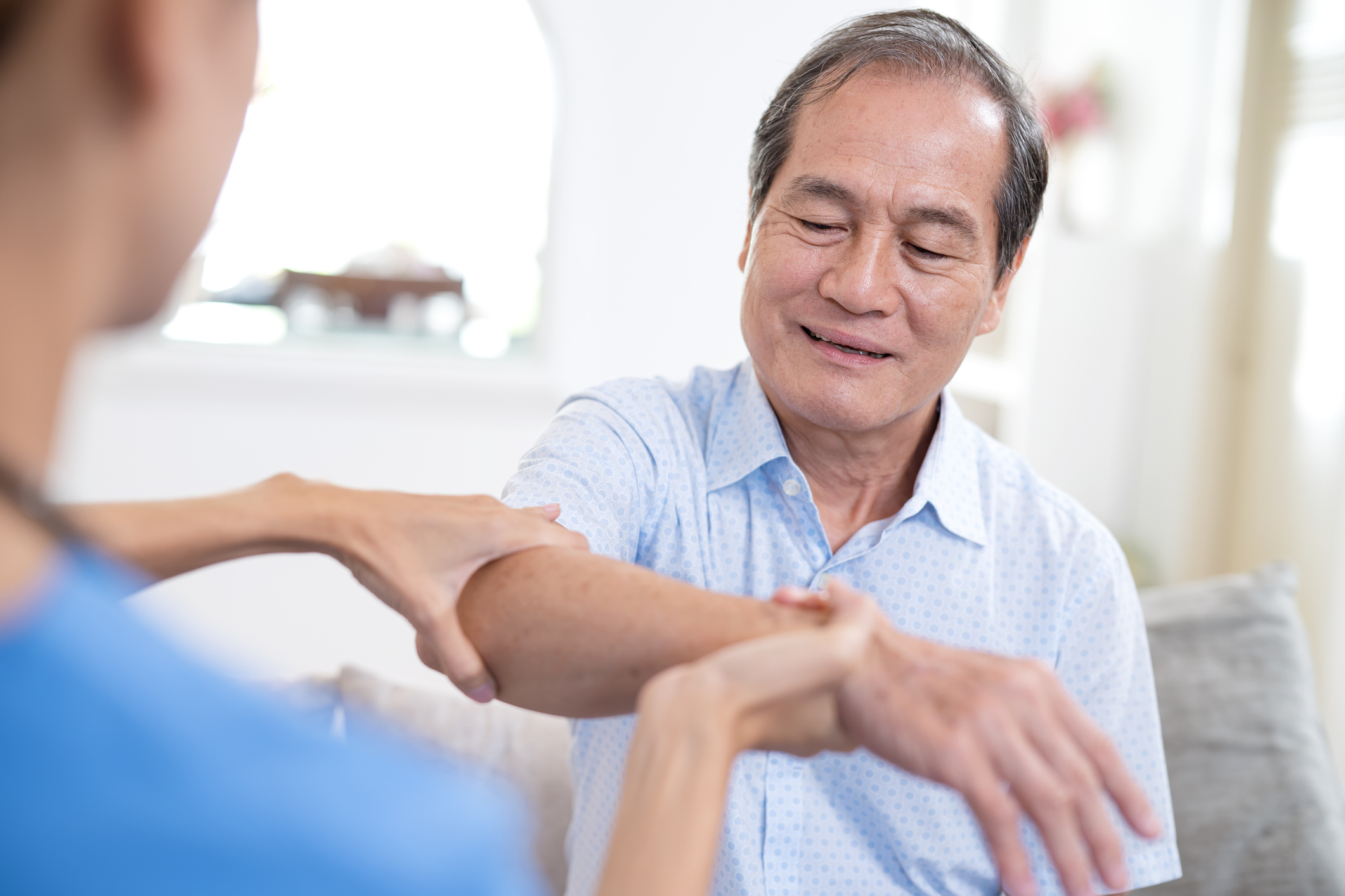 Geriatric Physical Therapy for Older Adults to Maintaining Independence in Performing Daily Activities. Home nurse helping a senior Asian man with massage of his hand and physical therapy to reduce myasthenia gravis disease at home.