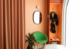 Corner of a orange themed doctors office with a cubicle for hanging clothes, a green chair and orange curtains