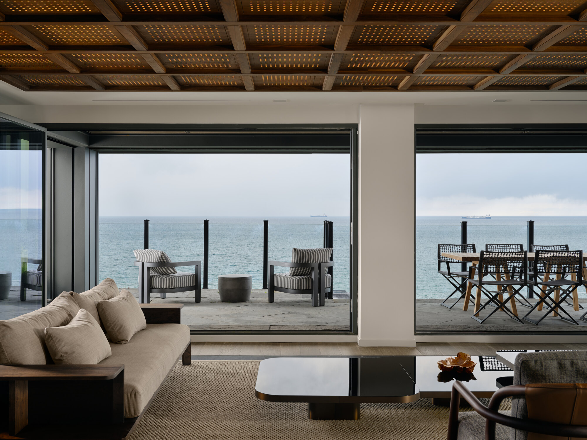 modern living room with lattice coffered ceiling, view out to balcony and lake beyond, beige sofas, black glass coffee table