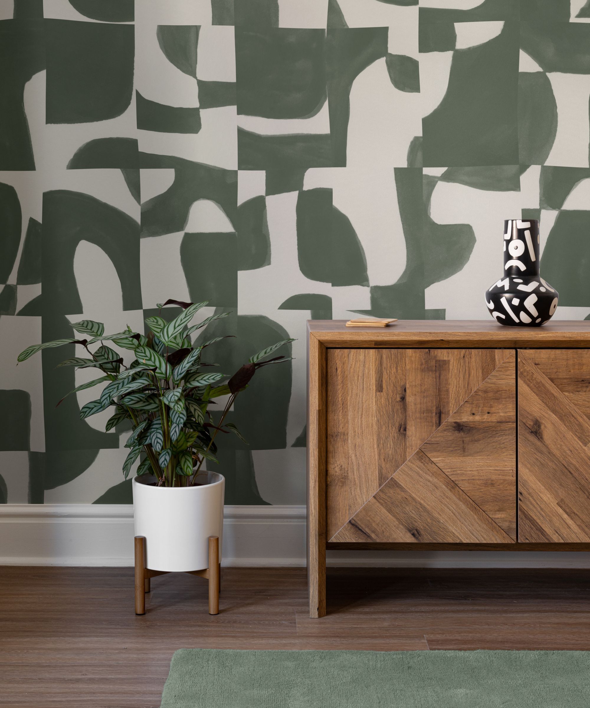 A modern space with large abstract green-and-white patterned wallpaper. A wooden sideboard, black-and-white vase, and potted plant complete the look.