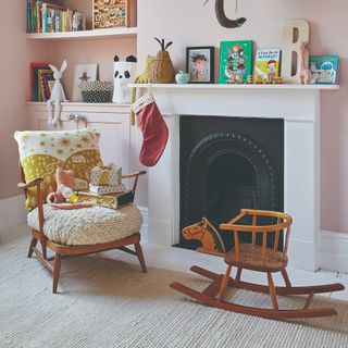 A pink-painted children's room with a fireplace mantelpiece and built-in alcove storage displaying toys, books and decor with a light beige rug covering white floorboards