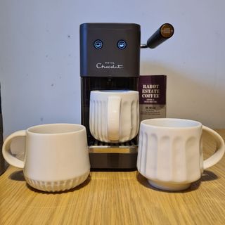 Hotel Chocolat Podster behind white mugs on wooden table