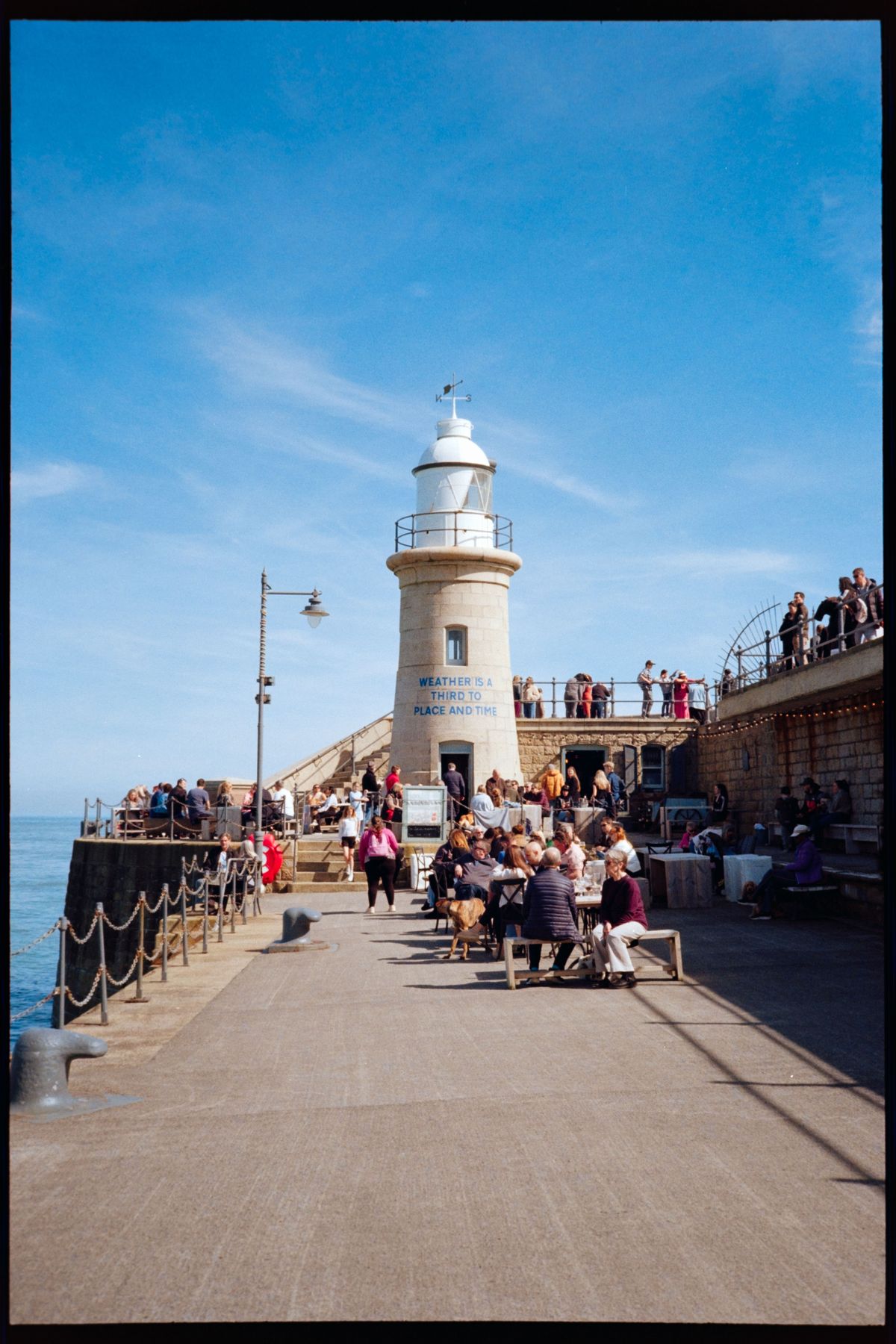 A white lighthouse on a busy pier with people sitting outside under a clear blue sky.