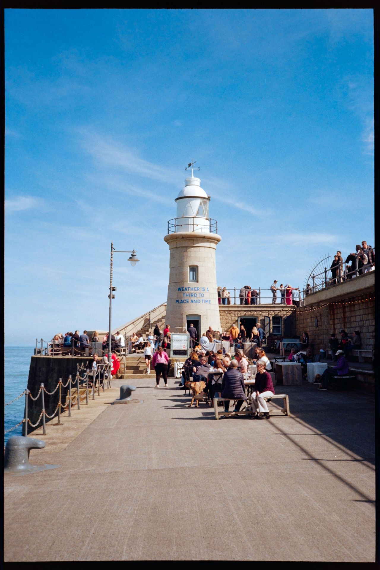 A white lighthouse on a busy pier with people sitting outside under a clear blue sky.
