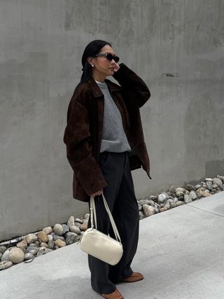 Fashion influencer Dawn Tan wearing a chic fall outfit consisting of a gray sweater, relaxed trousers, a vintage brown suede jacket, suede loafers, sunglasses, and a cream handbag.