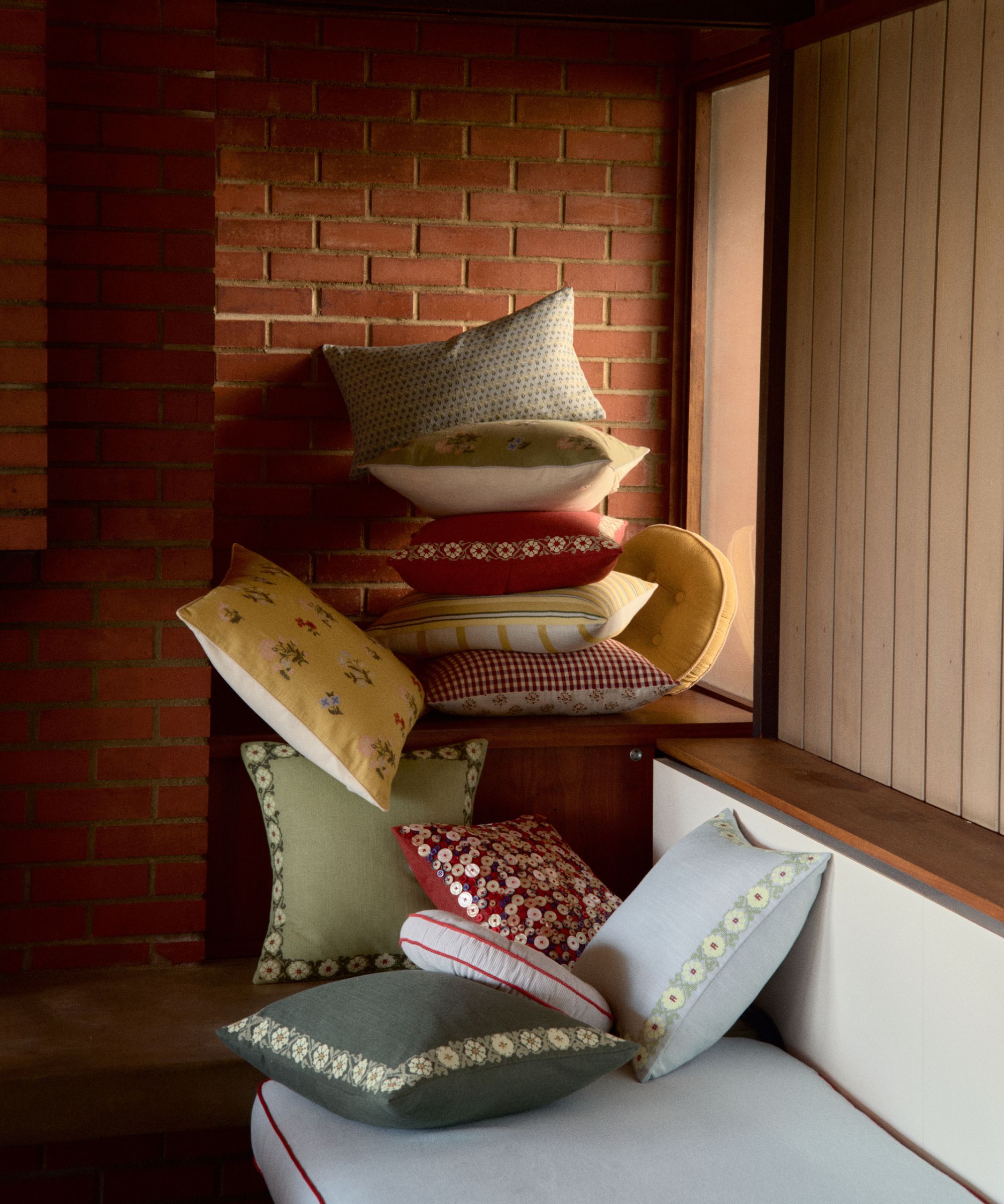 Image of several light blue, red, yellow, and green throw pillow stacked on the corner of a light blue bench seat and wooden ledge. There is a brick wall behind it.