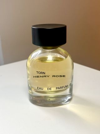 Henry rose torn perfume with large black circular cap on white desk against a grey wall