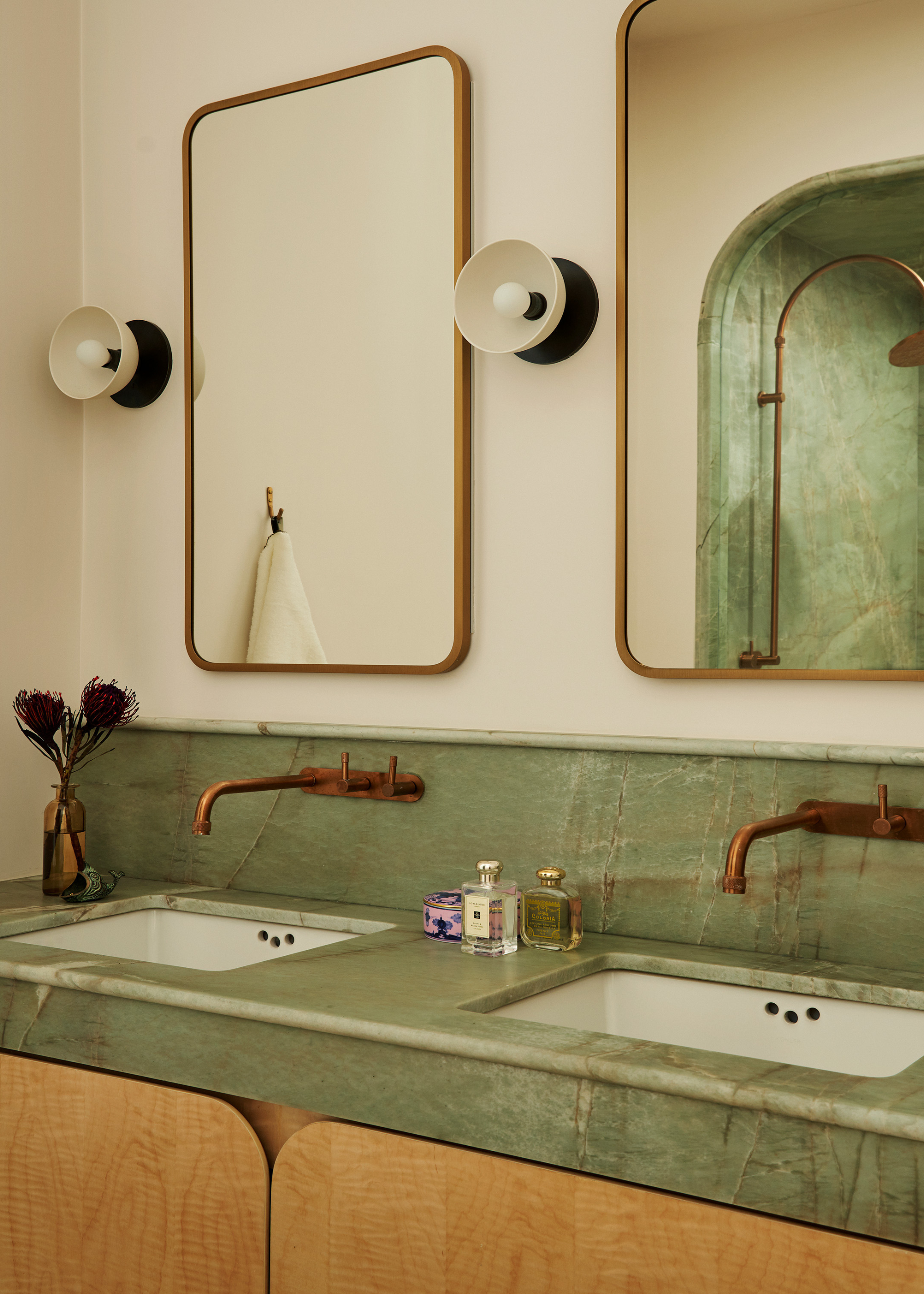 A bathroom with his and hers stations, a green marble counter, gold-rimmed mirrors, black and white sconces, brass hardware, and wooden cabinets