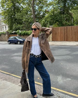 Lucy wears a brown leather jacket, white t-shirt, studded belt, dark blue jeans, black ballet flats, and a tote bag.