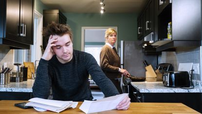 A young man or recent college grad is sitting at the kitchen table looking at bills or job applications. His mother looks on concerned in the background.