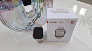 Huawei Watch Fit 3 står bredvid sin tillhörande förpackning på ett vitt bord.