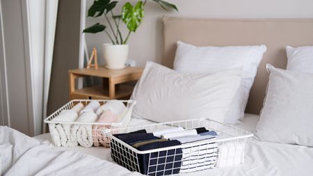 Two white wire baskets filled with folded clothes on a bed with white bedding. A wooden nightstand in the background with a white pot plant with long green stems and leaves. 