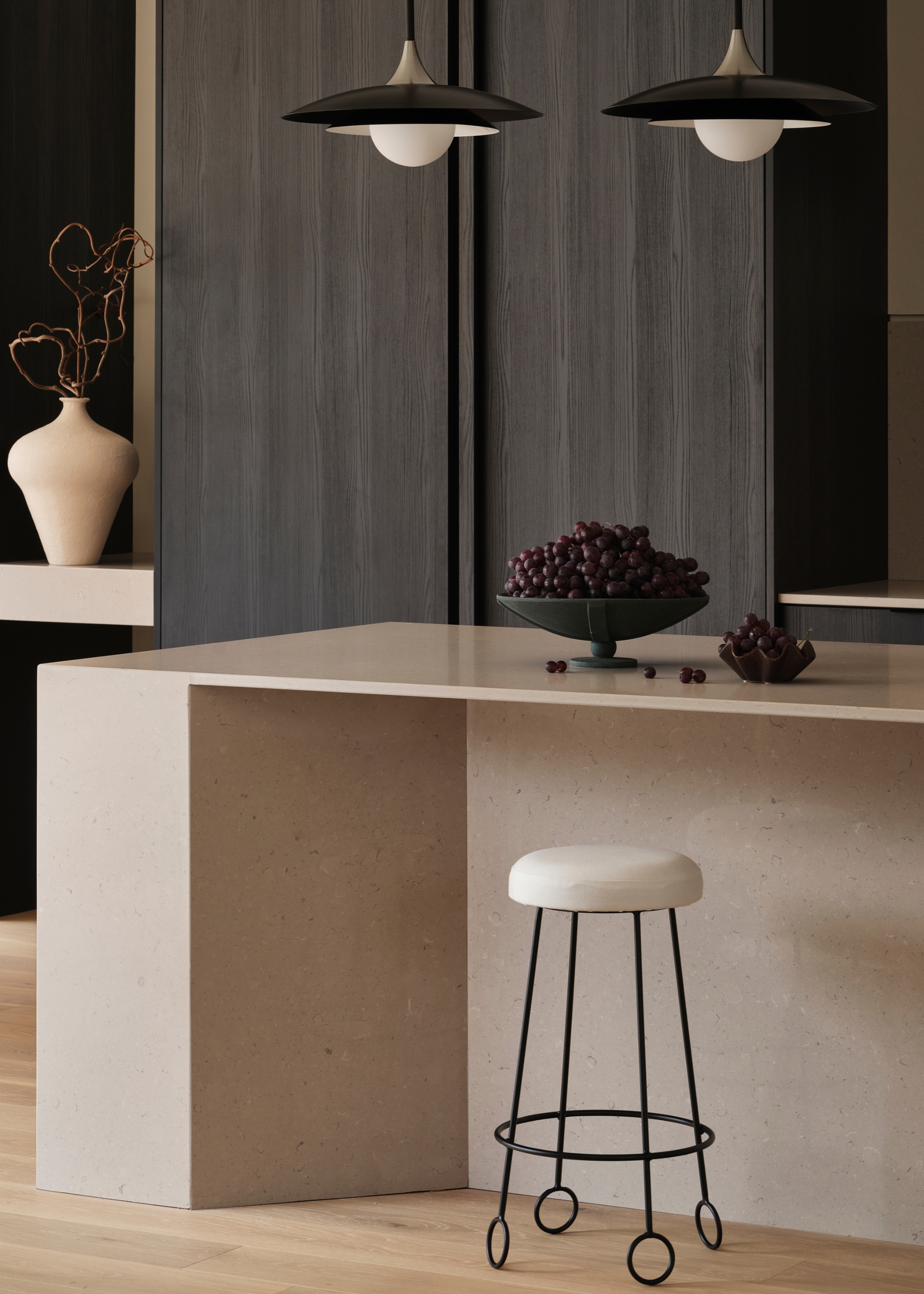A minimalist kitchen with dark wood cupboards, a pair of pendant lights, a marble island, a black metal stool, a bowl of grapes, and a vase of stems