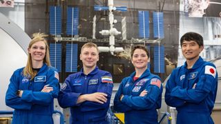 two men and two women in blue flight suits stand in front of a model of the international space station