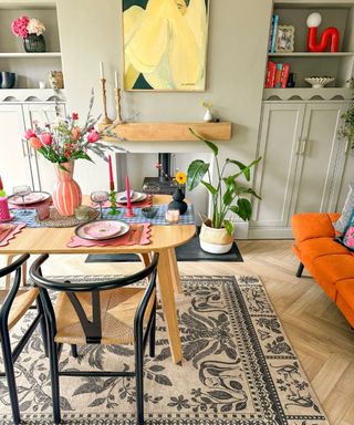 A dining area with a wooden table with a pink flower vase, black chairs around it, a black and white floral rug, and a sage green wall with a butter yellow art print on it