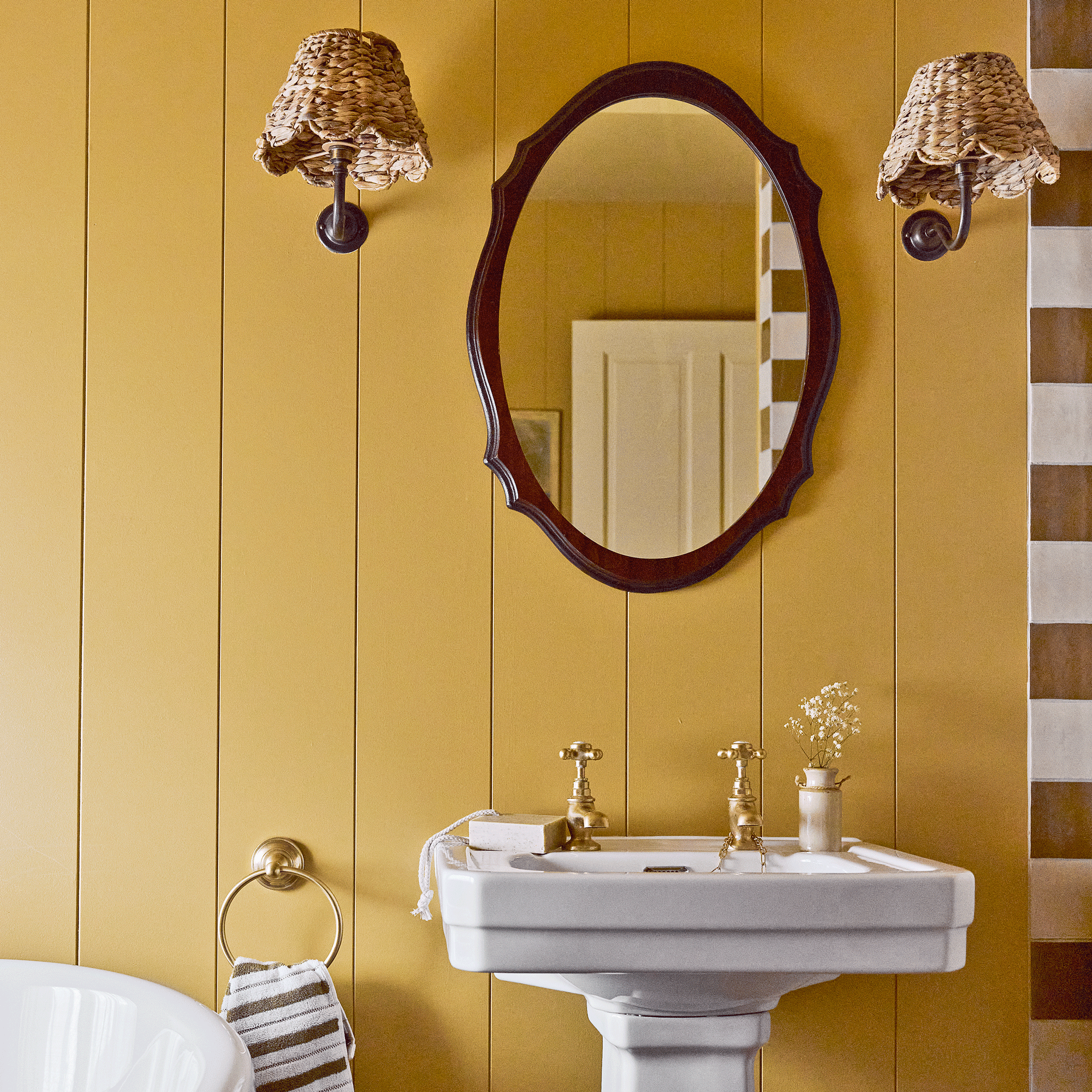 a yellow panelled bathroom with oval mirror and matching wicker wall lights either side