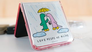 Samsung Galaxy Z Flip 7 in coralred in a cute case that says Love Keeps Us Alive and shows two dinosaurs sharing an umbrella
