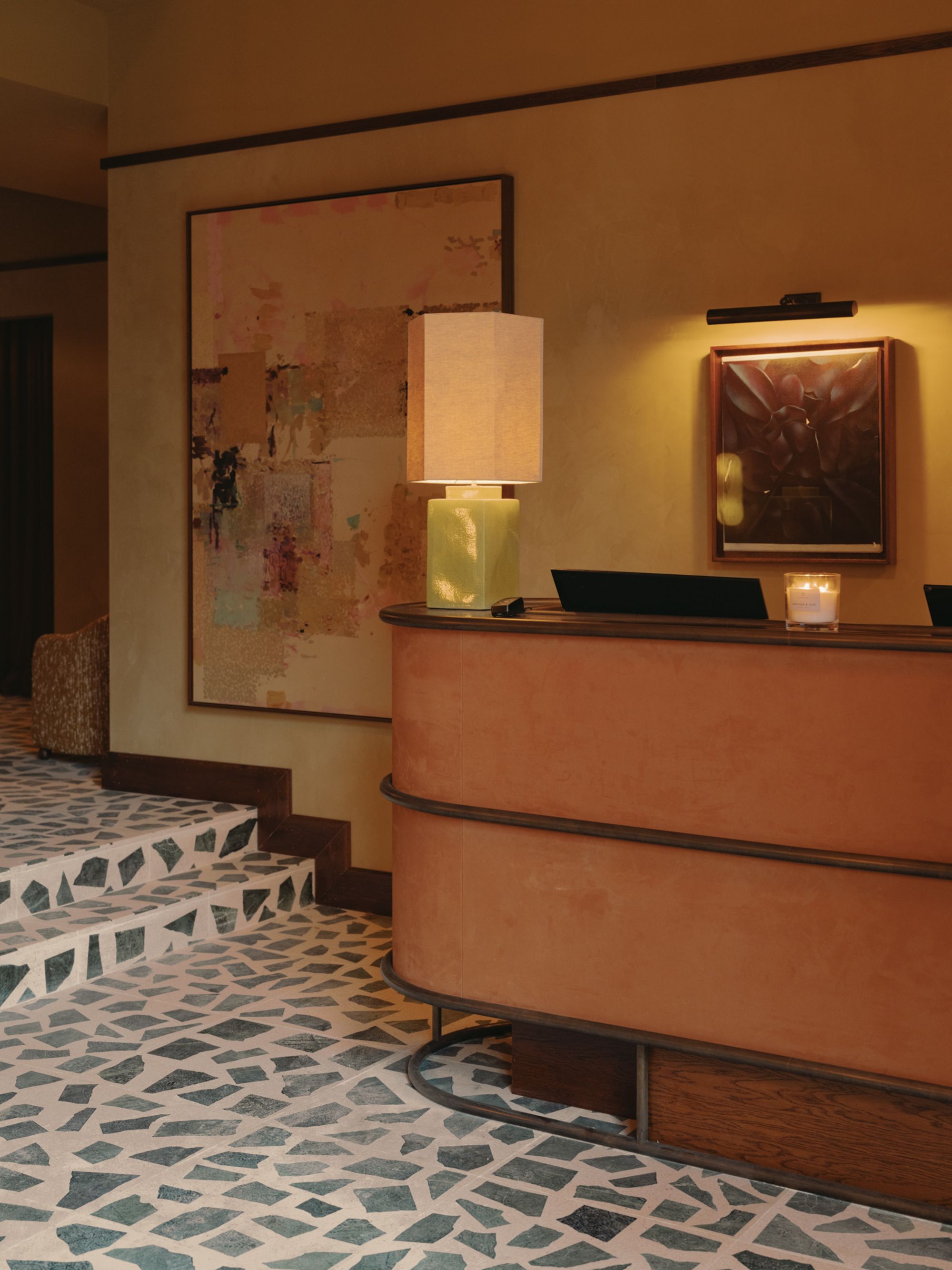 reception at soho house manchester with orange curved desk, terrazzo floors, steps, a large artwork, a table lamp on desk and candle burning