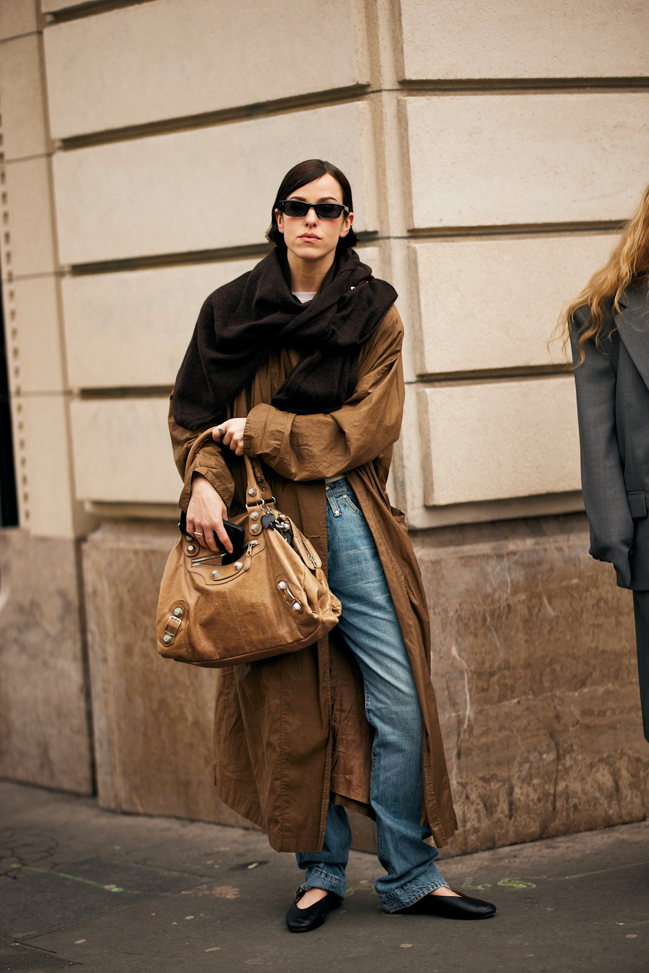 fashion week attendee wears sunglasses, wrap scarf, trench coat, balenciaga bag, jeans, and black ballet flats